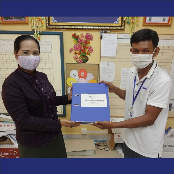 A man hands a blue folder of TukTuk worksheets to a woman in a face mask at an education office
