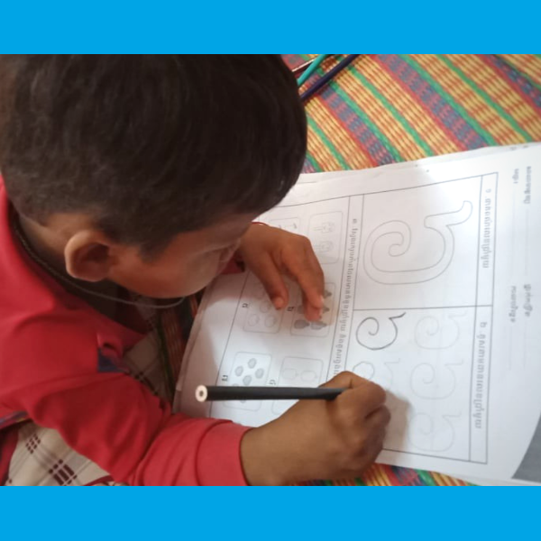 A child in a red shirt writes a Khmer letter on a TukTuk worksheet at home