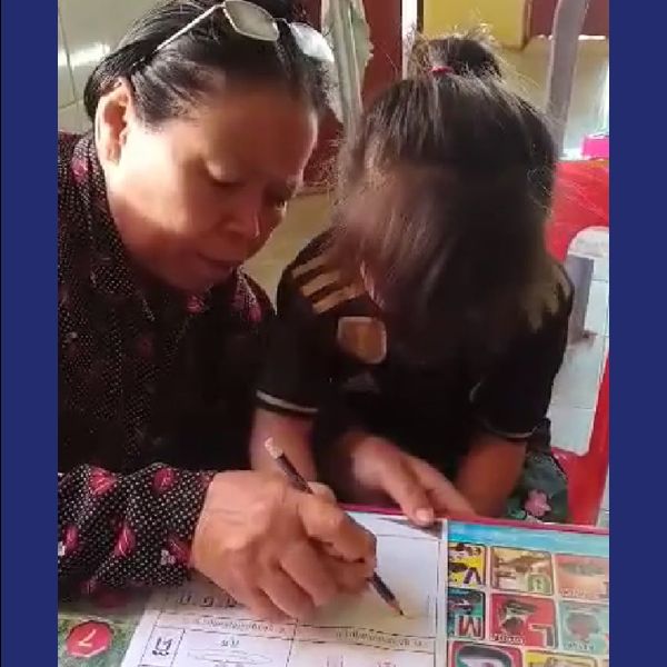 An older teacher helps a young child fill in a TukTuk worksheet at a table