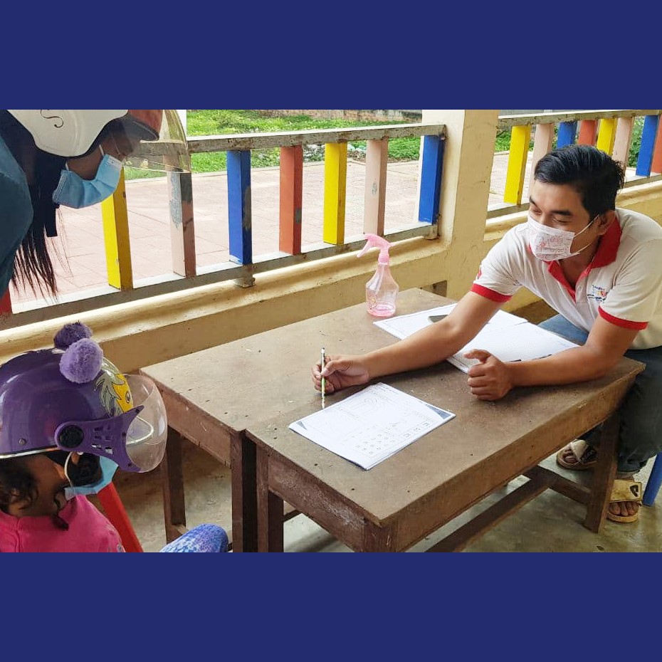 A TukTuk staff member in a mask writes at a table on a school veranda while a child waits