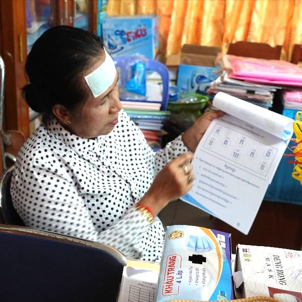 An older teacher in her school office holds up a TukTuk worksheet with counting exercises
