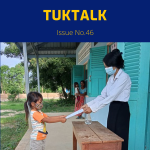 A teacher in a face mask hands a worksheet to a young girl at school