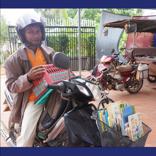 A man in a helmet on a motorbike carries a basket of picture books and a crate of worksheets