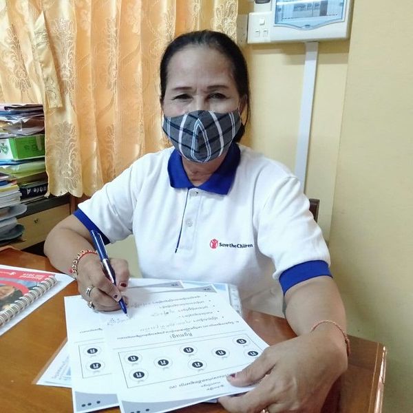 A teacher in a face mask sits at her desk and fills in a TukTuk worksheet