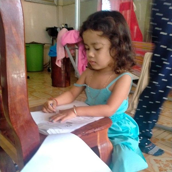 A young girl sits on a wooden chair and works on a TukTuk worksheet at home