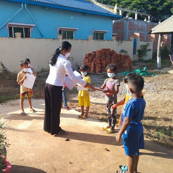 A teacher in a face mask hands worksheets to children outside a school building