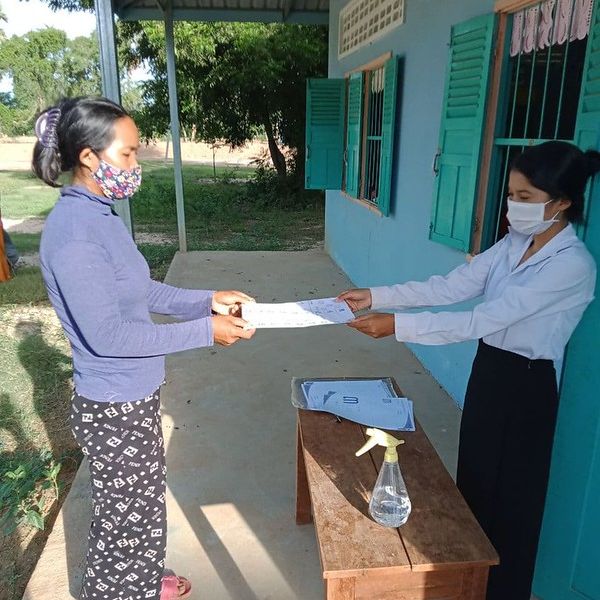 A woman in a face mask collects a worksheet from a teacher on a school veranda