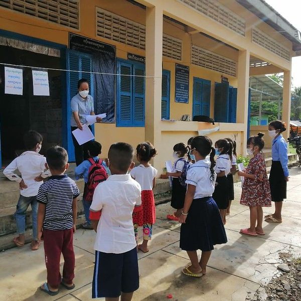 Children in school uniform queue outside a classroom while a teacher in a mask holds worksheets