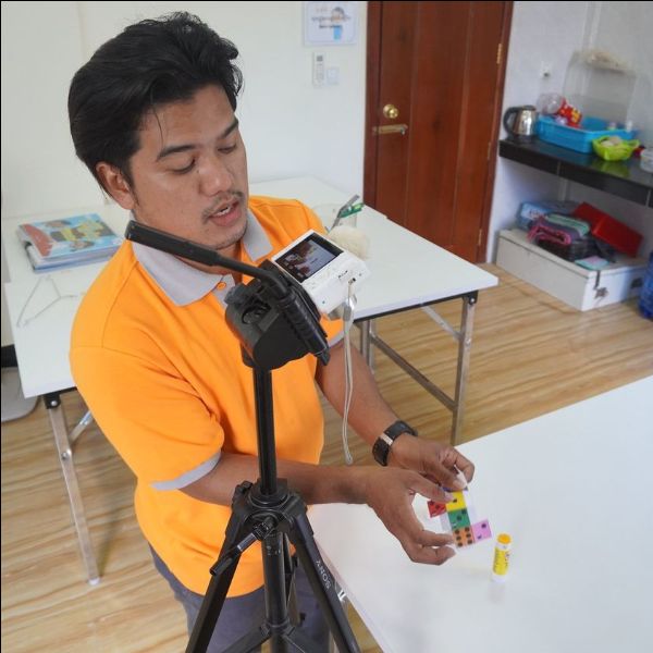 A TukTuk staff member in an orange shirt films a lesson video with a phone on a tripod