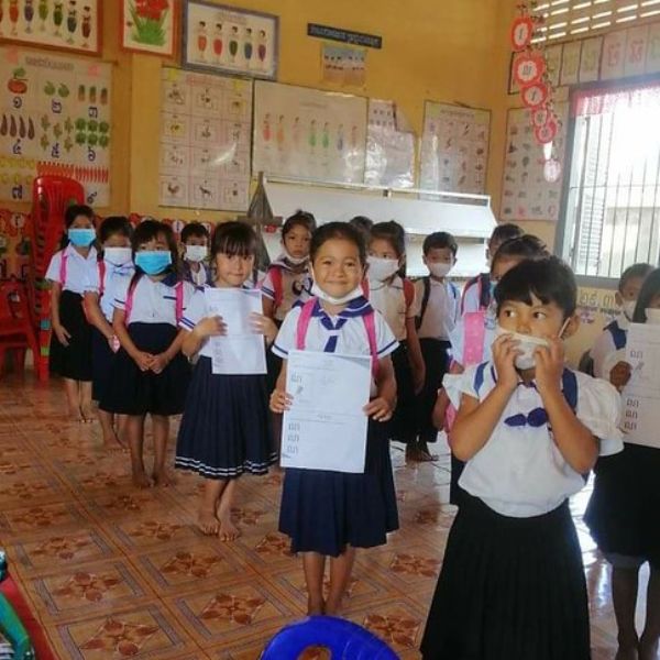 Kindergarten children in school uniform and face masks stand in a classroom holding their worksheets