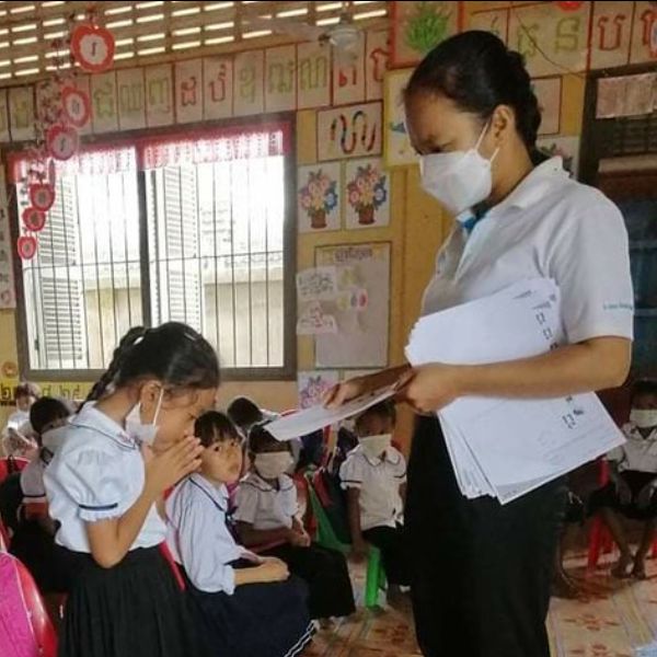 A teacher in a mask hands out worksheets to children sitting in a kindergarten classroom