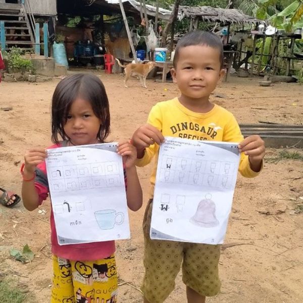 Two small children stand outside a house, each holding up a TukTuk worksheet