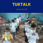 Masked kindergarten children walk in a classroom holding their worksheets
