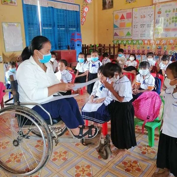 A teacher in a wheelchair shows a worksheet to a child in a crowded kindergarten classroom