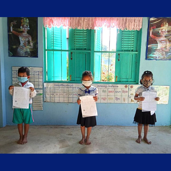 Three children stand barefoot in a classroom, each holding up a worksheet