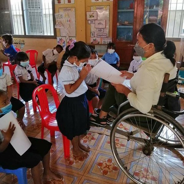 A teacher in a wheelchair reads from a worksheet to masked children sitting in a classroom