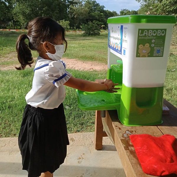 A girl in a face mask washes her hands at a green Happy Tap hand wash stand outdoors