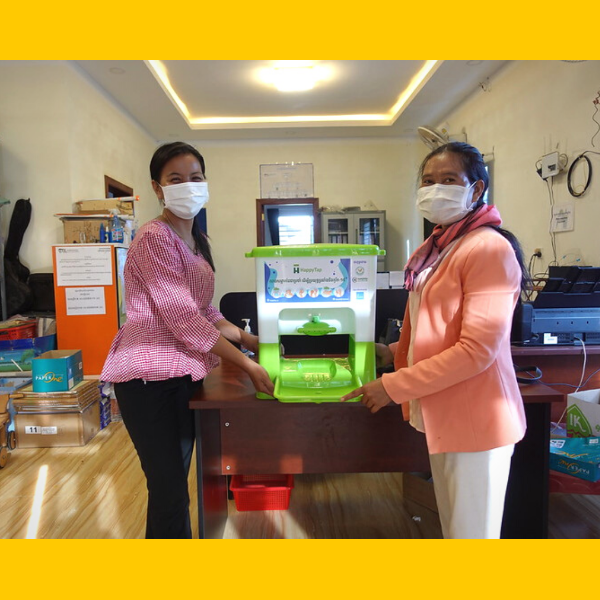 Two women in face masks hold a green hand wash stand on a desk in a school office