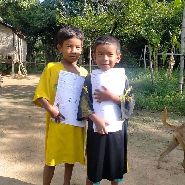 Two children stand outside a house, each holding a TukTuk worksheet
