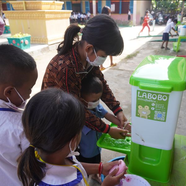 A teacher helps young children wash their hands at a green hand wash stand in a school yard