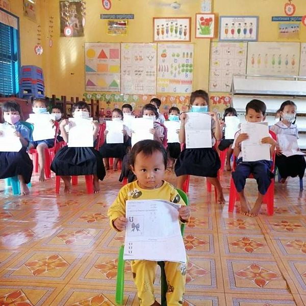 Preschool children in face masks sit on red chairs holding up their worksheets while a toddler sits in front