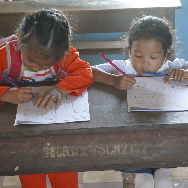 Two girls write carefully on TukTuk worksheets at a wooden school desk
