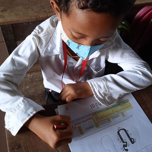 A boy in school uniform and a face mask writes on a Khmer worksheet