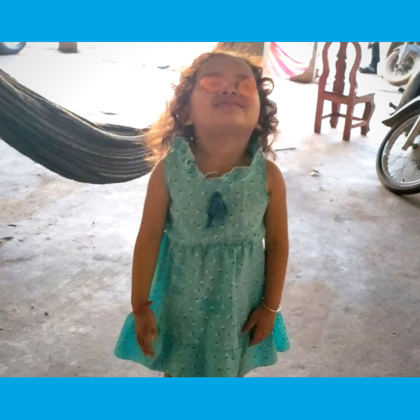 A little girl in a teal dress stands with her eyes closed in front of a hammock