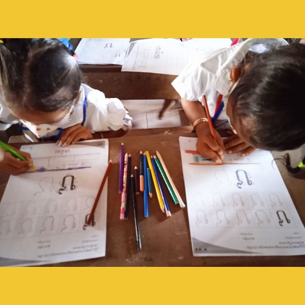 Two children in school uniform colour in Khmer letter worksheets with coloured pencils