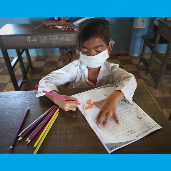 A girl in a face mask colours a worksheet next to a set of coloured pencils