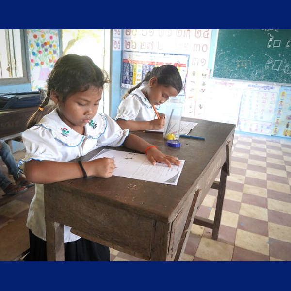 Two girls in school uniform write on worksheets at wooden desks in a classroom