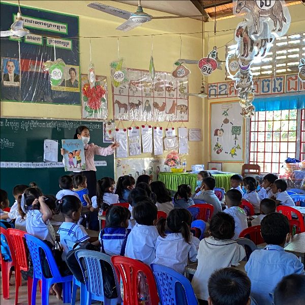 A teacher reads a picture book to a classroom of children sitting on plastic chairs