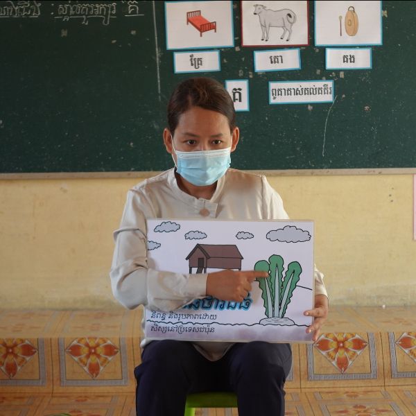 A teacher in a face mask holds a picture card of a house and a turnip plant
