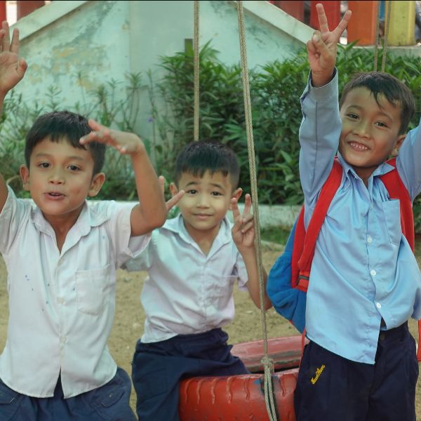 Three boys in school uniform wave and cheer next to a tyre swing in a school yard