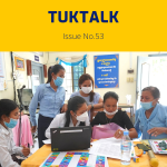 Teachers and TukTuk staff gather around a laptop and colourful worksheets at a workshop table