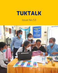 Teachers and TukTuk staff gather around a laptop and colourful worksheets at a workshop table