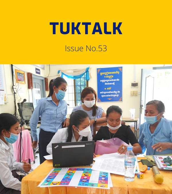 Teachers and TukTuk staff gather around a laptop and colourful worksheets at a workshop table
