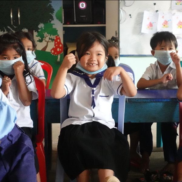 A smiling girl in school uniform clenches her fists playfully in a classroom of masked children