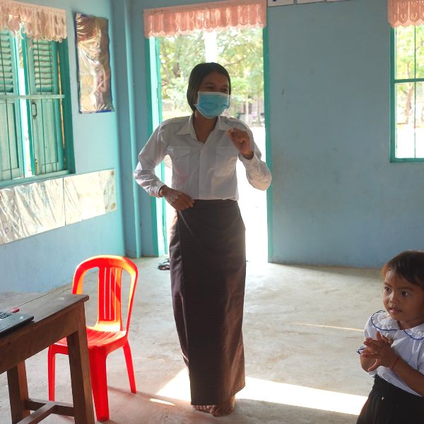 A teacher in a face mask walks across a classroom while a child watches from the edge