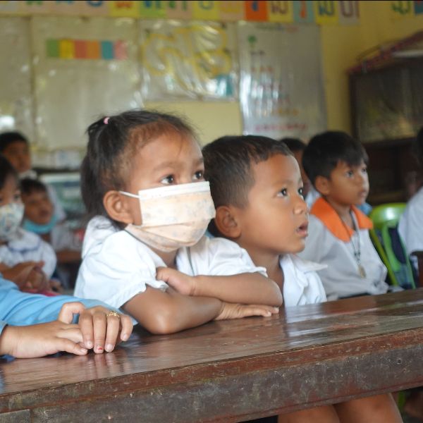 A girl in a face mask rests her arms on a wooden desk and listens in class