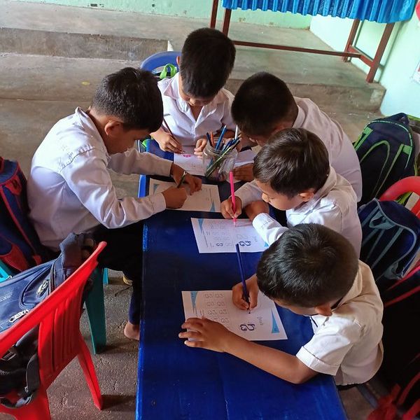 Boys in white shirts work on worksheets together around a blue table
