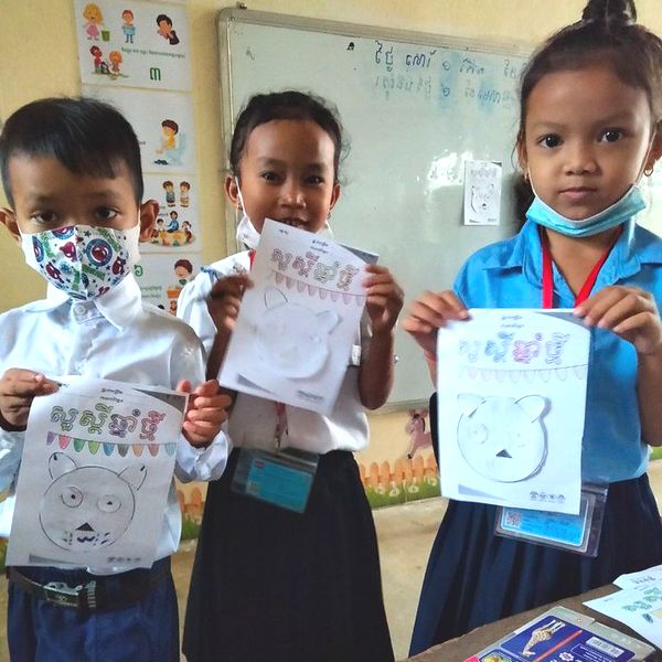 Three children hold up Khmer worksheets with animal mask drawings in a classroom