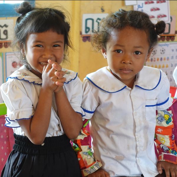 Two preschool girls in school uniform smile at the camera, one with her hands clasped