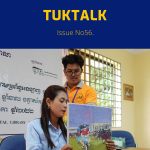 A TukTuk staff member and a teacher hold up a picture book at a desk under a Khmer banner