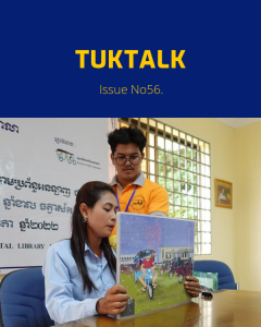 A TukTuk staff member and a teacher hold up a picture book at a desk under a Khmer banner