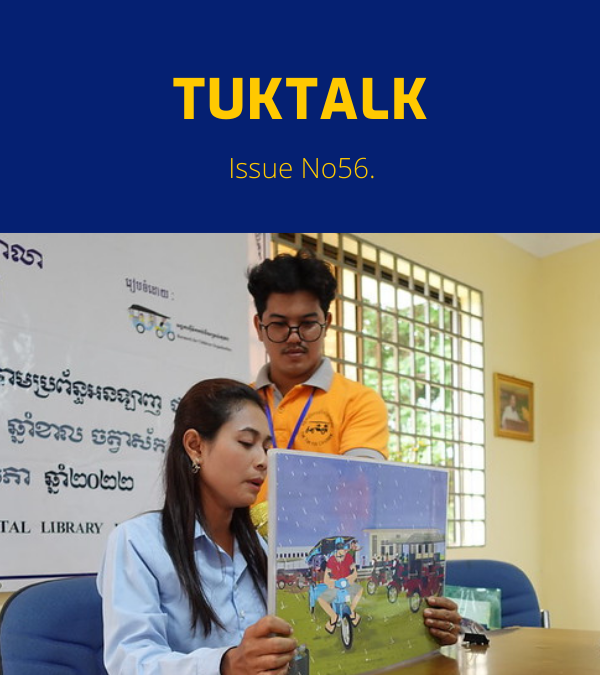 A TukTuk staff member and a teacher hold up a picture book at a desk under a Khmer banner