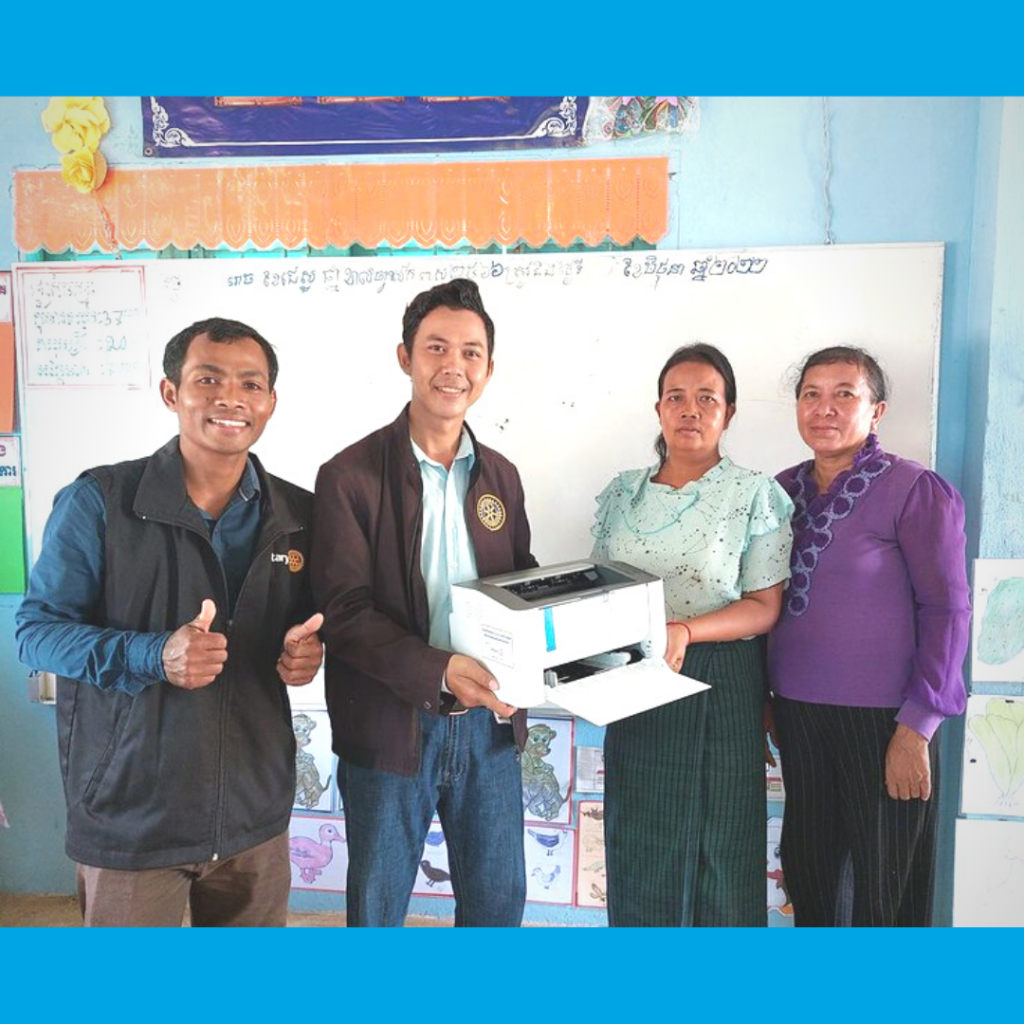 TukTuk staff hand a printer to two teachers in a classroom, one giving a thumbs up