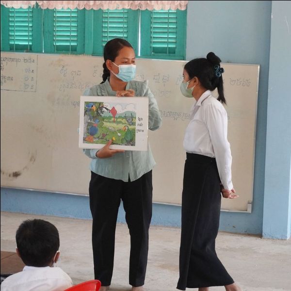 A teacher holds up a picture poster while a colleague stands beside her in a classroom