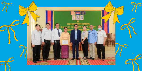 Education officials and TukTuk representatives pose with a certificate in a hall decorated with yellow ribbons