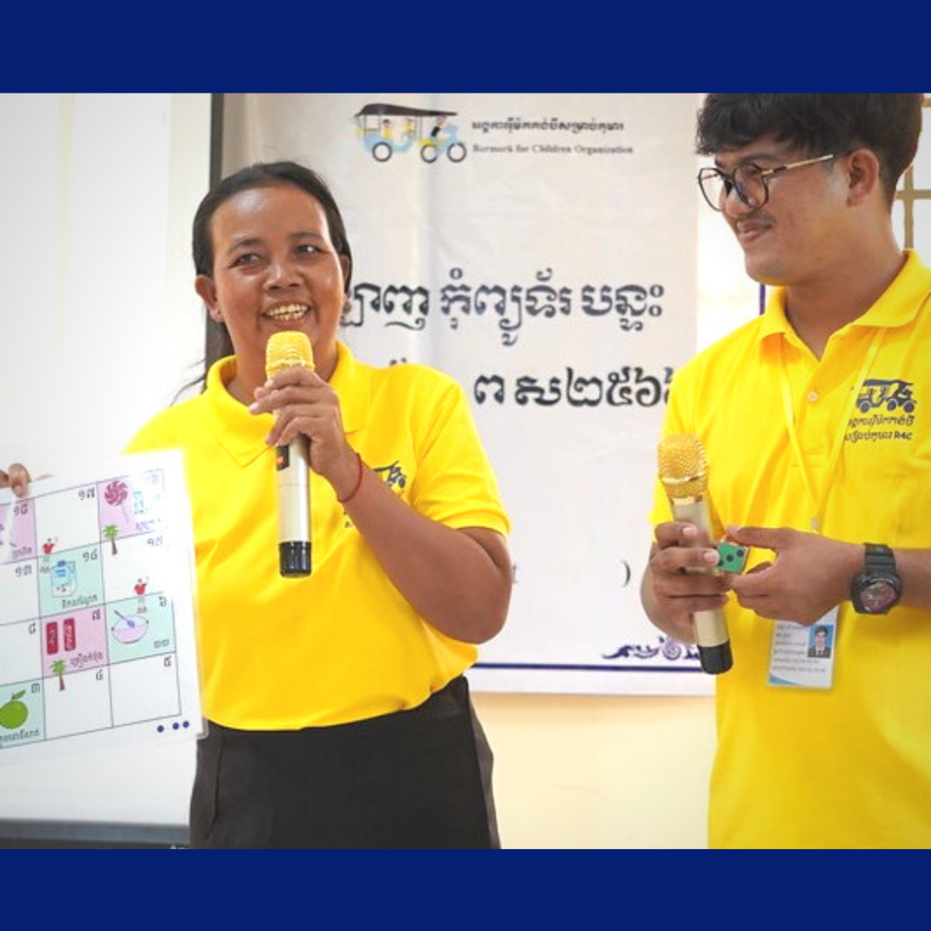 A woman in a yellow TukTuk shirt speaks into a microphone while holding a game board at the workshop